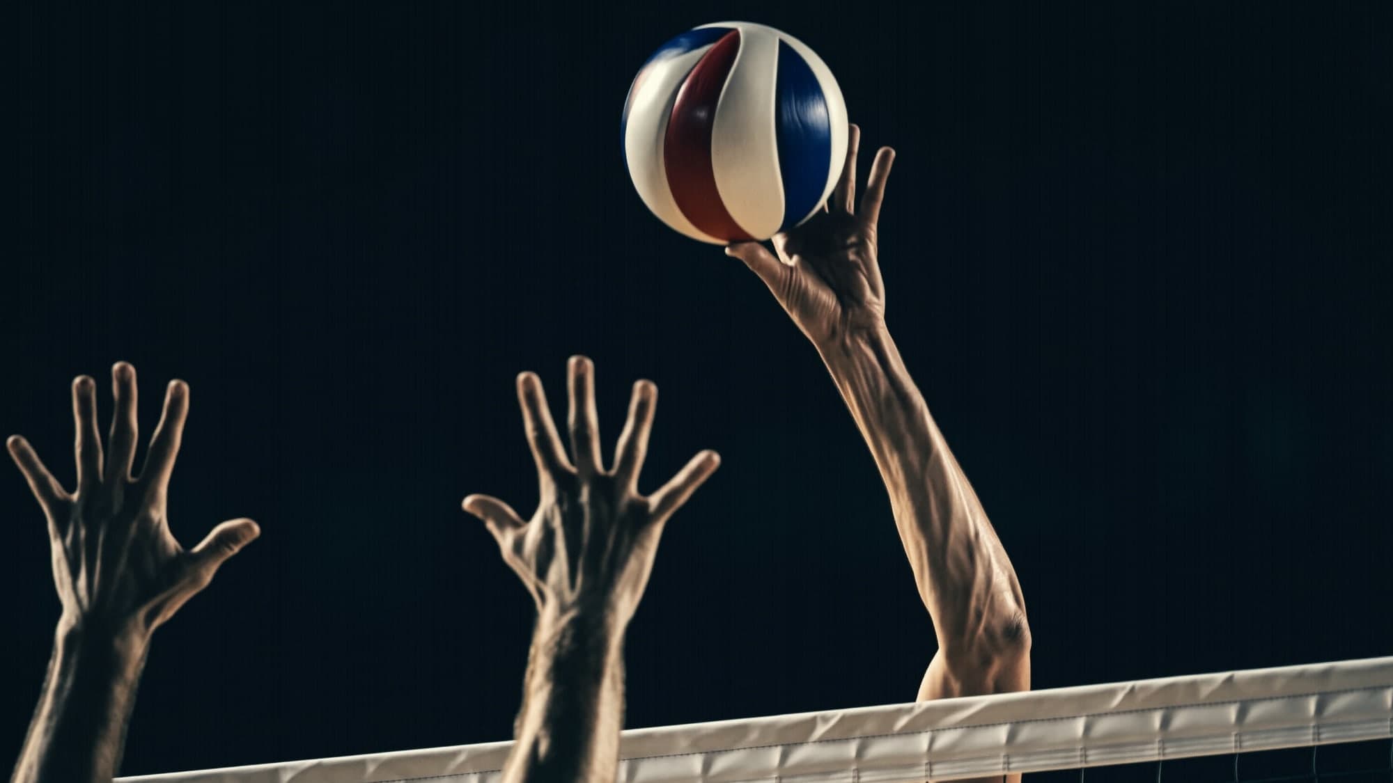 A volleyball compressing against an outstretched hand at the top of a spike, a blocker's hands rising opposite.