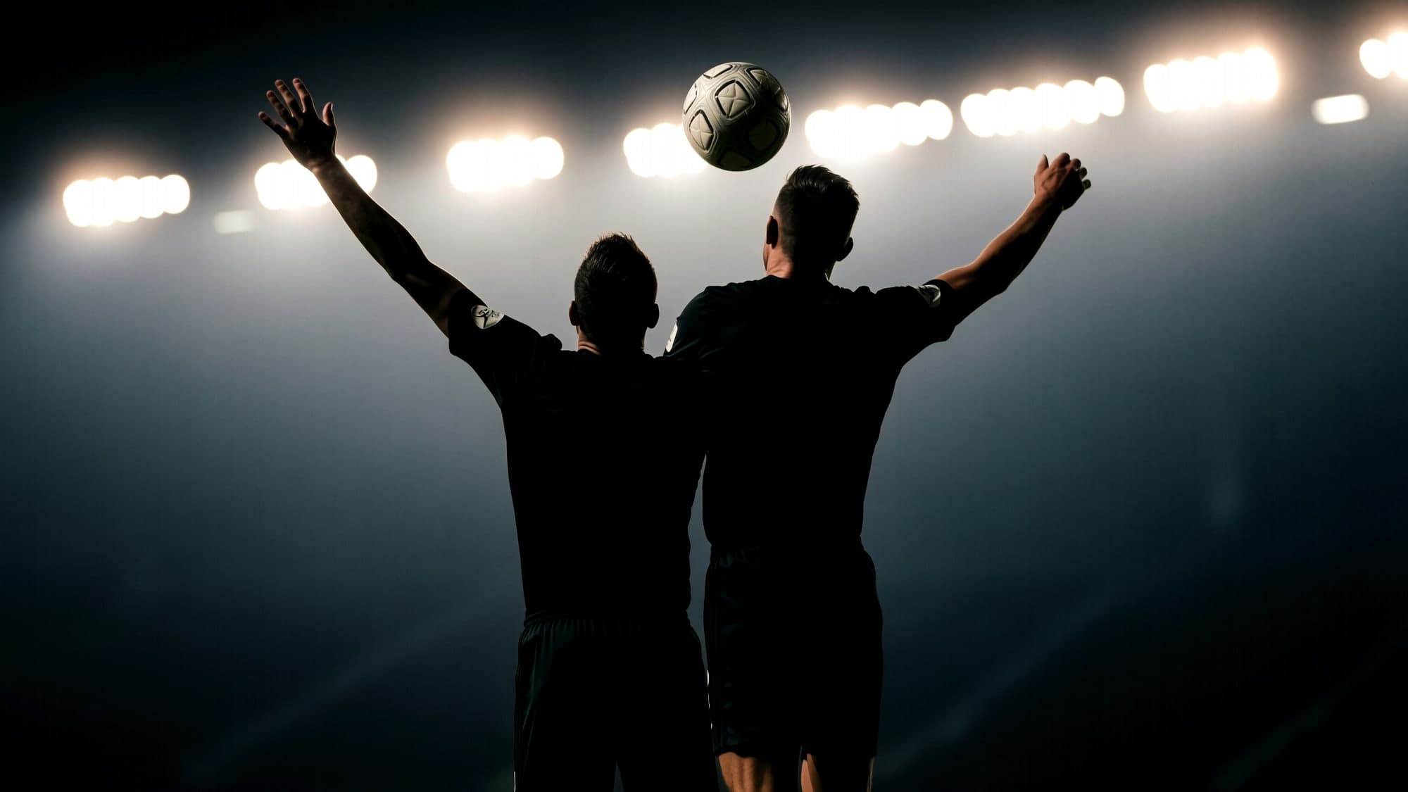 Two players seen from behind, arms raised, a ball above them against a bank of stadium floodlights.