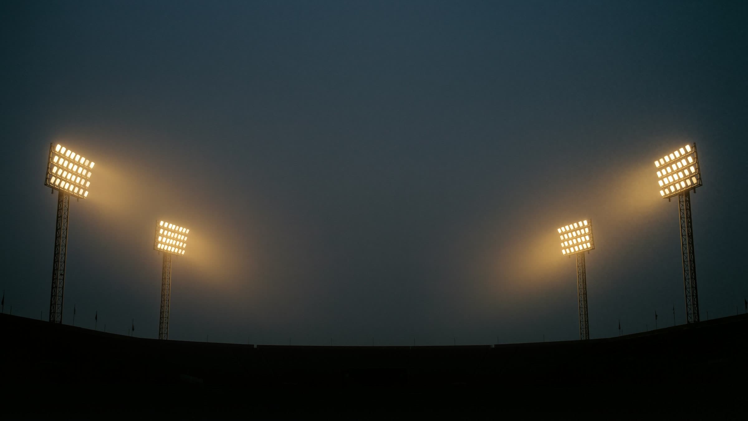 Stadium floodlight towers glowing through haze against a near-black dusk sky.