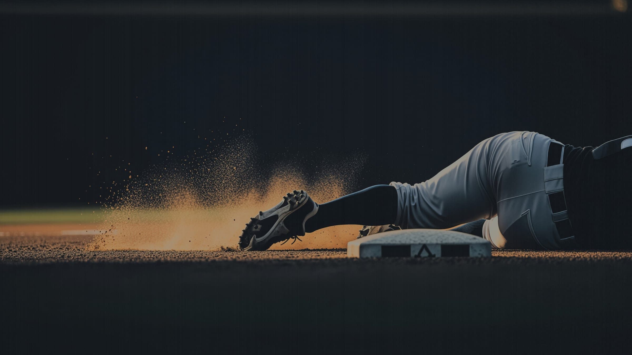 A baserunner sliding into a base at dusk, cleats first, throwing up a backlit spray of infield dust.
